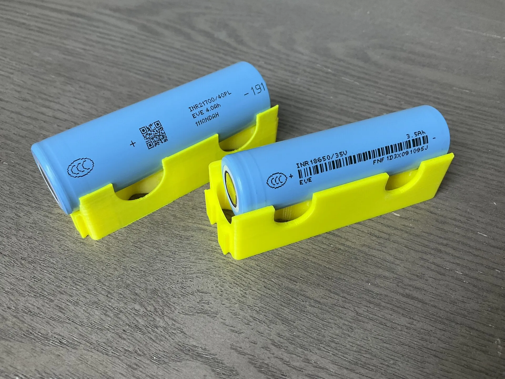 Deux cellules lithium-ion cylindriques bleues, une 18650 et une 21700 un peu plus grosse, posées sur des supports jaunes