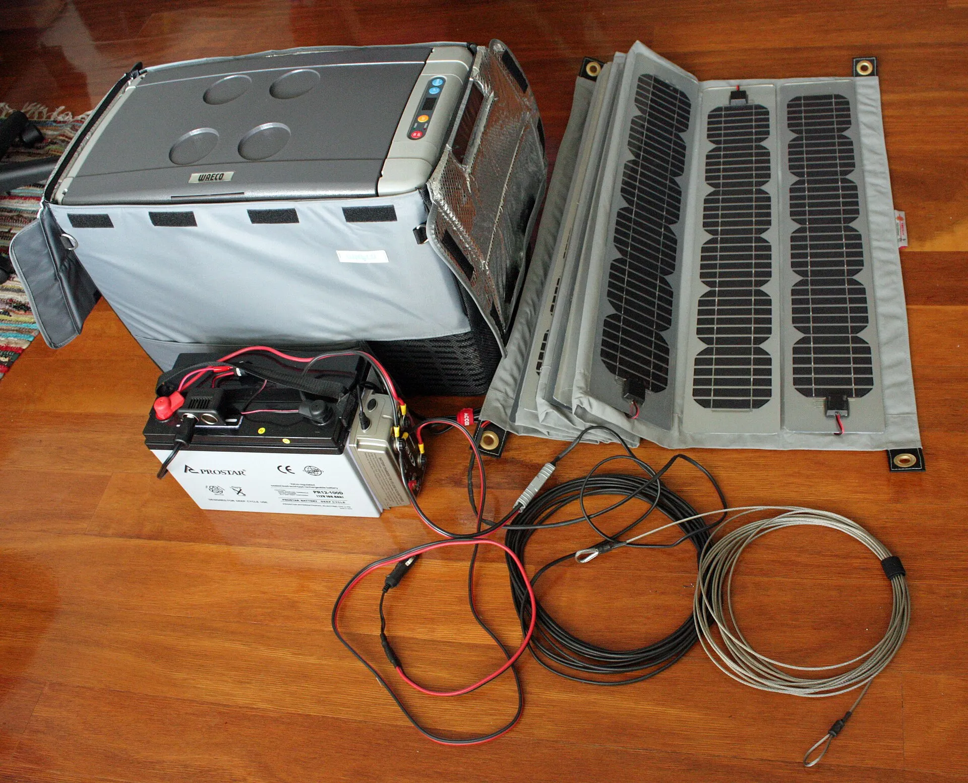 Équipement électrique d'un véhicule aménagé posé au sol : panneau solaire souple replié, batterie auxiliaire, réfrigérateur 12 V et câbles