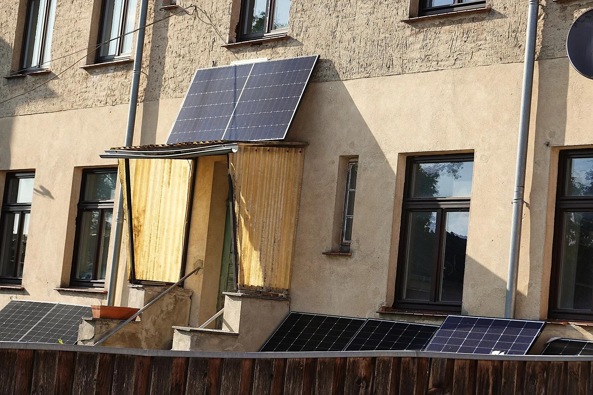 Façade d'immeuble avec plusieurs petits kits solaires : un panneau sur un auvent et d'autres posés contre la clôture