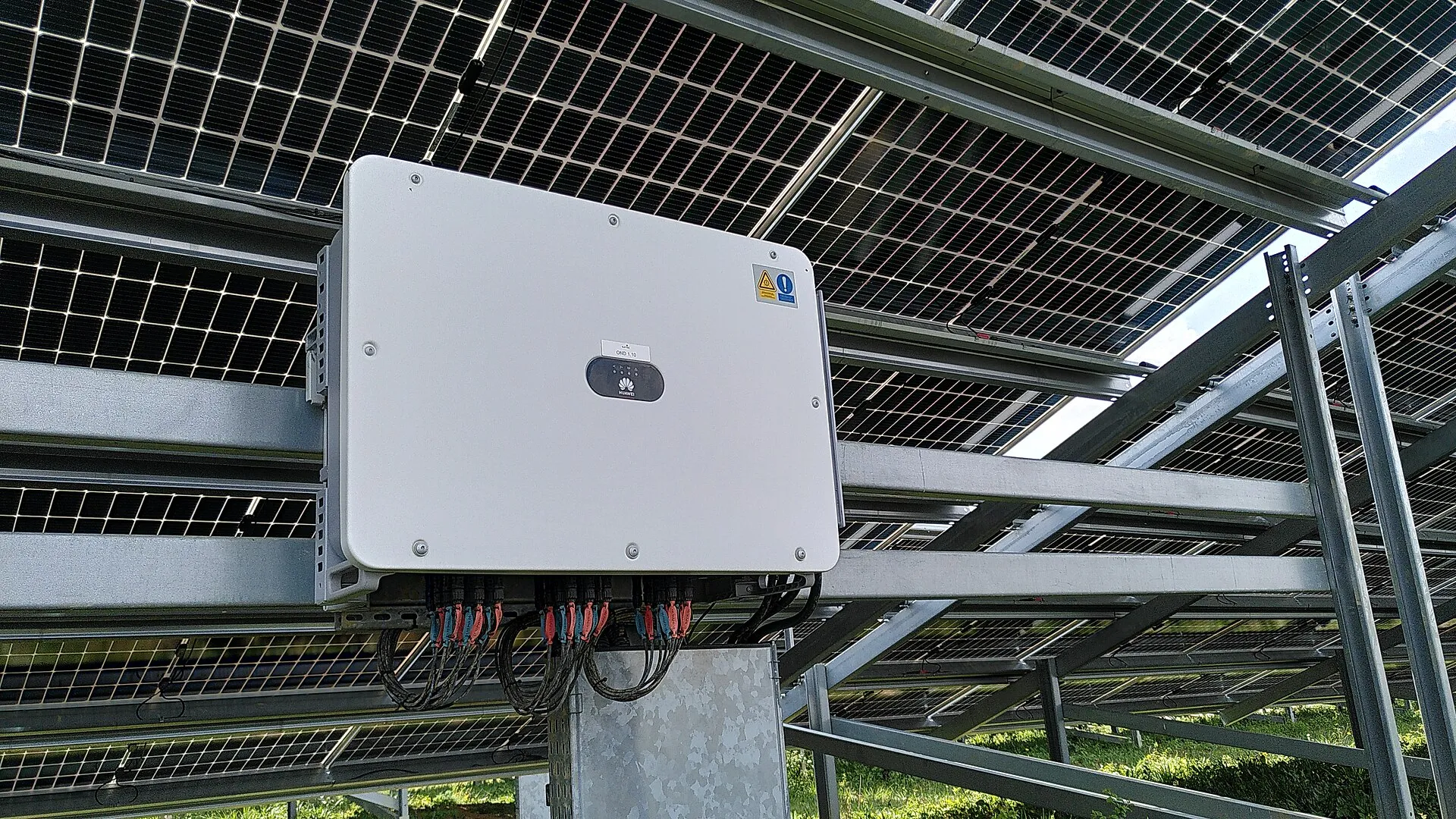 Onduleur string gris fixé sous une rangée de panneaux solaires, avec ses câbles branchés en dessous