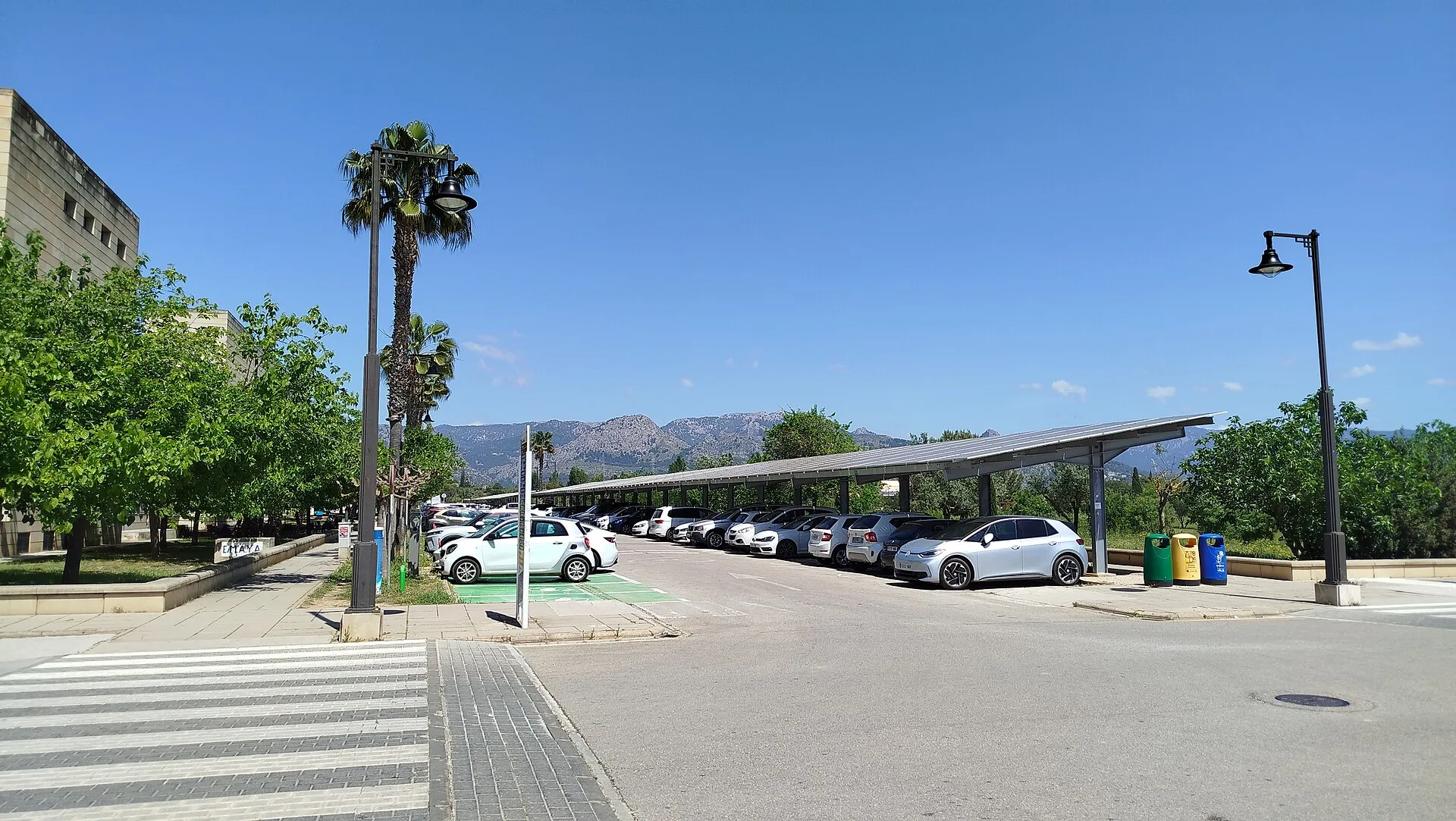 Longue pergola couverte de panneaux photovoltaïques au-dessus d'un parking, voitures garées en épi dessous, palmiers et montagnes au loin