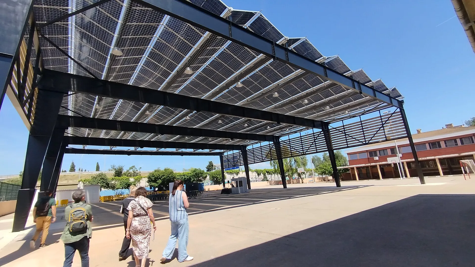 Pergola solaire dans une cour d'école : des panneaux photovoltaïques posés sur une structure métallique font de l'ombre aux passants